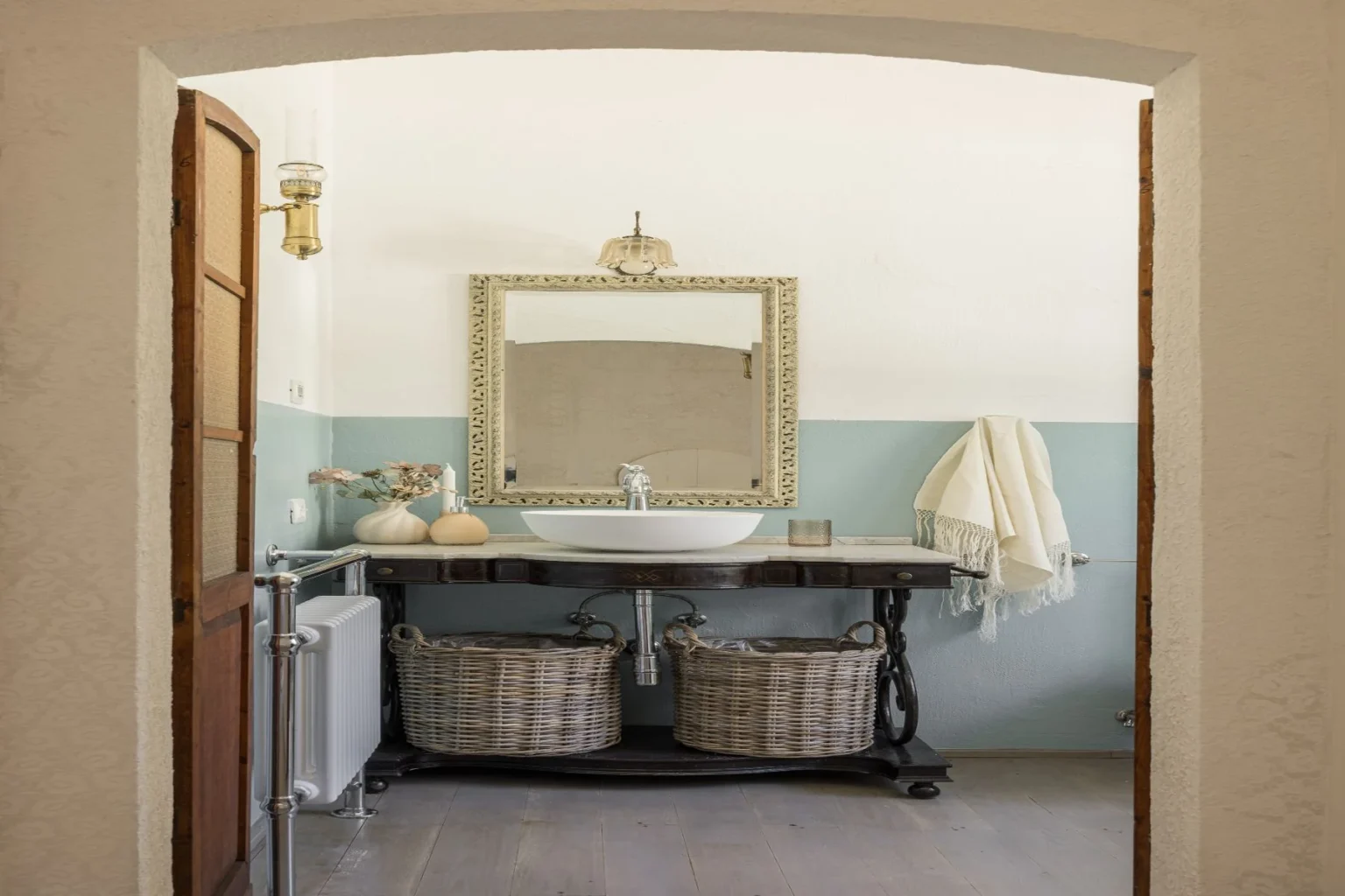 Elegant bathroom interior featuring a vintage wooden vanity with a round sink, decorative mirror, and stylish lighting. The space is accented by soft blue wall paint, a towel hanging on the side, and woven baskets for storage, creating a serene and inviting atmosphere. | Only on SopranoVillas