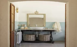 Elegant bathroom interior featuring a vintage wooden vanity with a round sink, decorative mirror, and stylish lighting. The space is accented by soft blue wall paint, a towel hanging on the side, and woven baskets for storage, creating a serene and inviting atmosphere.