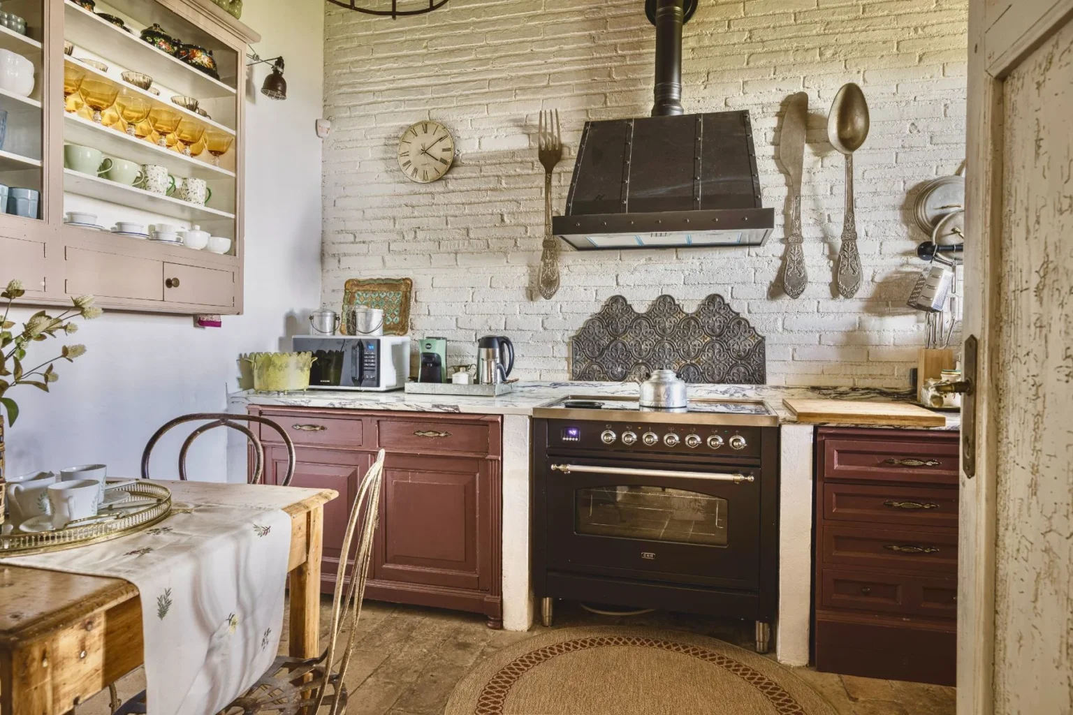 Cozy kitchen interior featuring rustic wooden furniture, vintage decor, and modern appliances. The space includes a black oven, a microwave, and a kettle, complemented by a marble countertop and a decorative backsplash. Natural light enhances the inviting atmosphere, with a dining table set for tea and elegant glassware displayed on open shelves. | Only on SopranoVillas