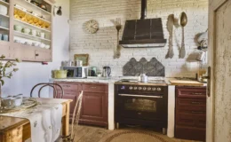 Cozy kitchen interior featuring rustic wooden furniture, vintage decor, and modern appliances. The space includes a black oven, a microwave, and a kettle, complemented by a marble countertop and a decorative backsplash. Natural light enhances the inviting atmosphere, with a dining table set for tea and elegant glassware displayed on open shelves.