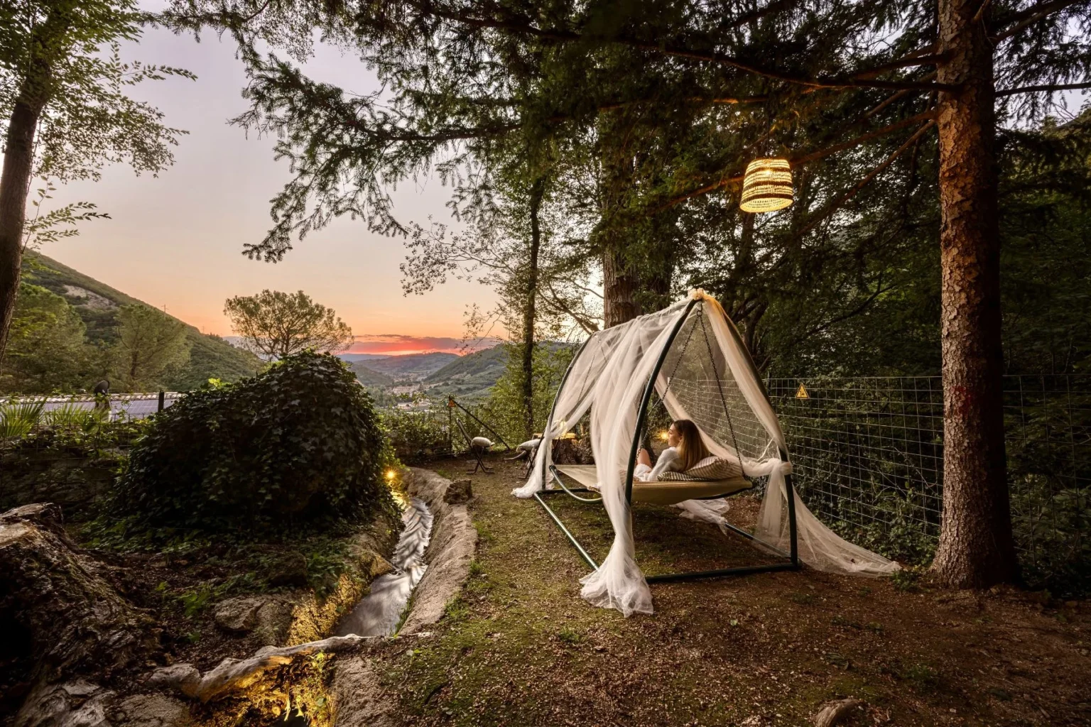 Cozy outdoor hammock nestled in a serene forest setting, featuring a woman relaxing under a canopy of sheer fabric with a stunning sunset view over the mountains. Lush greenery surrounds a gentle stream, enhancing the tranquil atmosphere. | Only on SopranoVillas