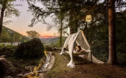 Cozy outdoor hammock nestled in a serene forest setting, featuring a woman relaxing under a canopy of sheer fabric with a stunning sunset view over the mountains. Lush greenery surrounds a gentle stream, enhancing the tranquil atmosphere.