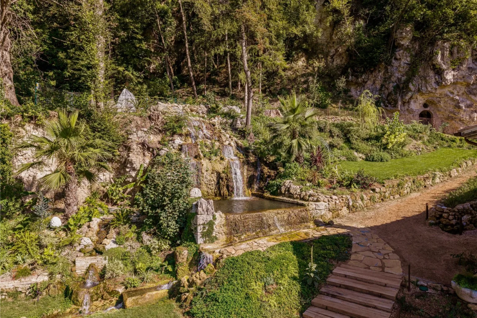 Lush garden landscape featuring a serene waterfall cascading into a clear pond, surrounded by vibrant greenery and tropical plants, with a stone pathway leading through the natural setting. | Only on SopranoVillas