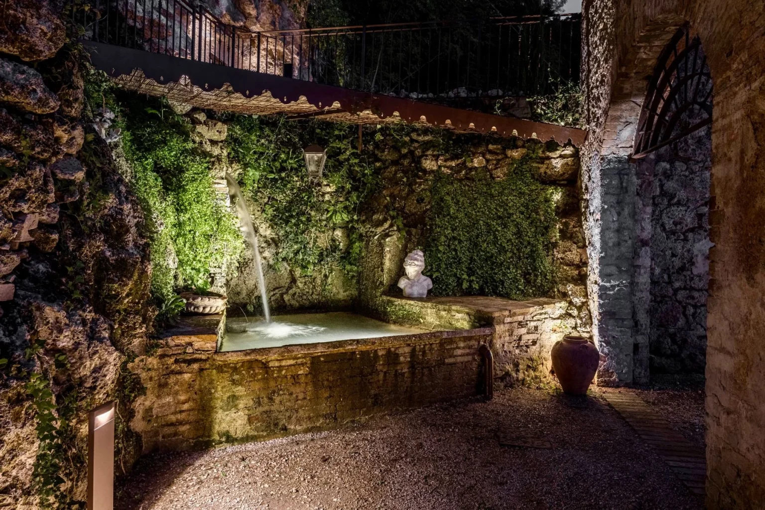 Serene garden water feature with cascading water, surrounded by lush green ivy and rustic stone walls, featuring a decorative statue and ambient lighting. Ideal for creating a tranquil outdoor retreat. | Only on SopranoVillas