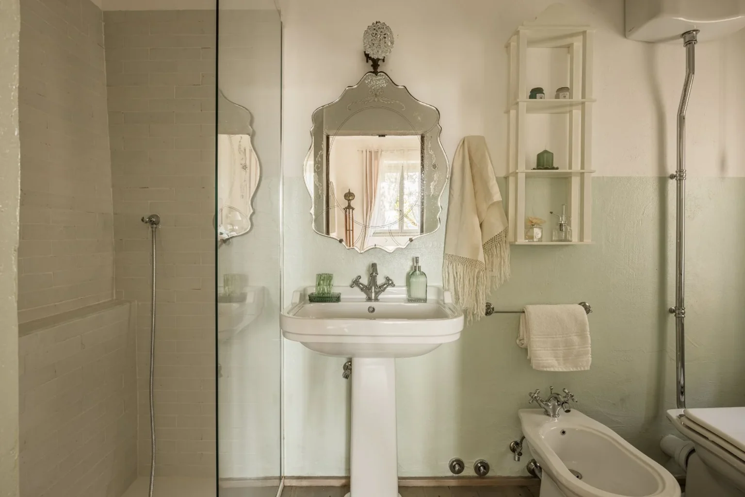 Modern bathroom featuring a glass shower, vintage-style mirror, and elegant sink. The walls are painted in soft green and white, complemented by decorative shelves holding toiletries. A towel hangs neatly beside the sink, creating a serene and stylish atmosphere. | Only on SopranoVillas