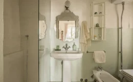 Modern bathroom featuring a glass shower, vintage-style mirror, and elegant sink. The walls are painted in soft green and white, complemented by decorative shelves holding toiletries. A towel hangs neatly beside the sink, creating a serene and stylish atmosphere.