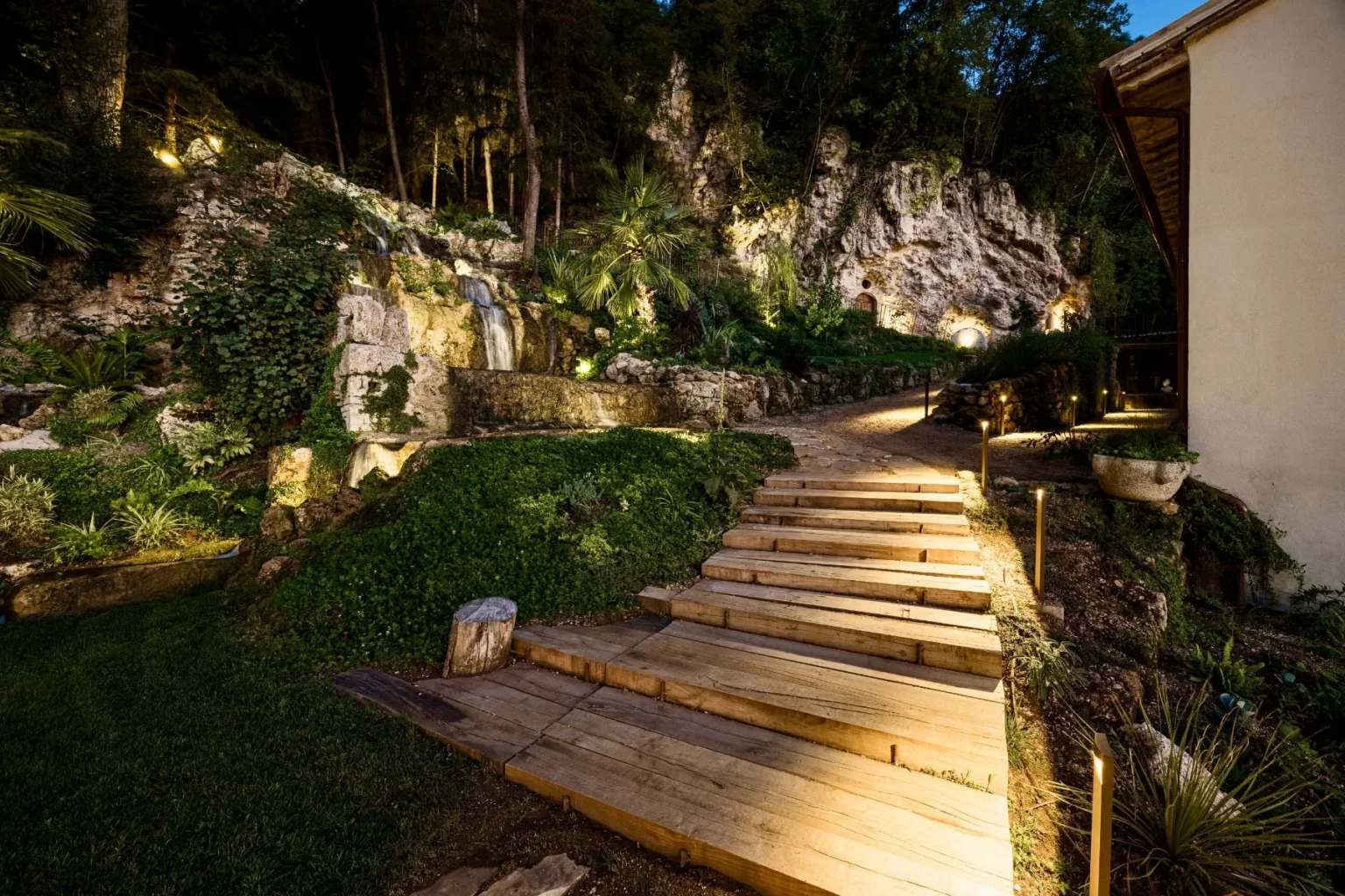 Wooden steps lead through a beautifully landscaped garden featuring a waterfall, lush greenery, and ambient lighting, set against a rocky backdrop at night. | Only on SopranoVillas