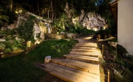 Wooden steps lead through a beautifully landscaped garden featuring a waterfall, lush greenery, and ambient lighting, set against a rocky backdrop at night.