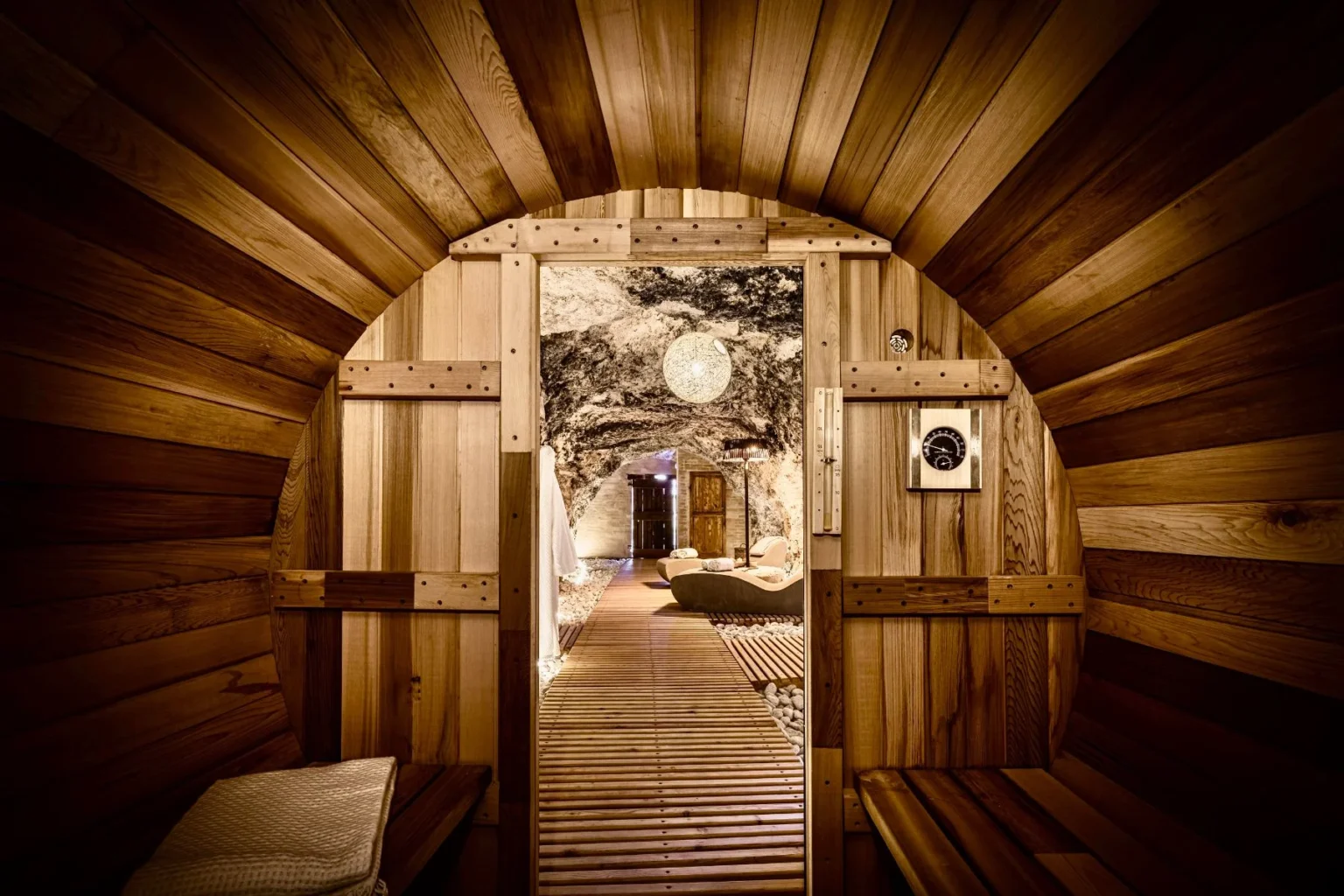 Interior view of a wooden sauna with a circular door, showcasing natural wood paneling and a serene pathway leading to a relaxation area with soft lighting and stone accents. | Only on SopranoVillas