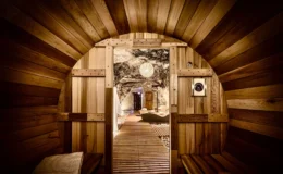 Interior view of a wooden sauna with a circular door, showcasing natural wood paneling and a serene pathway leading to a relaxation area with soft lighting and stone accents.