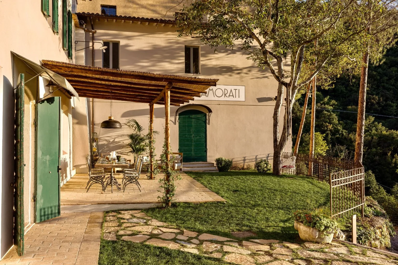Charming outdoor seating area at a rustic villa with green doors and a shaded pergola, surrounded by lush greenery and stone pathways, ideal for relaxation and dining. | Only on SopranoVillas