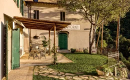 Charming outdoor seating area at a rustic villa with green doors and a shaded pergola, surrounded by lush greenery and stone pathways, ideal for relaxation and dining.