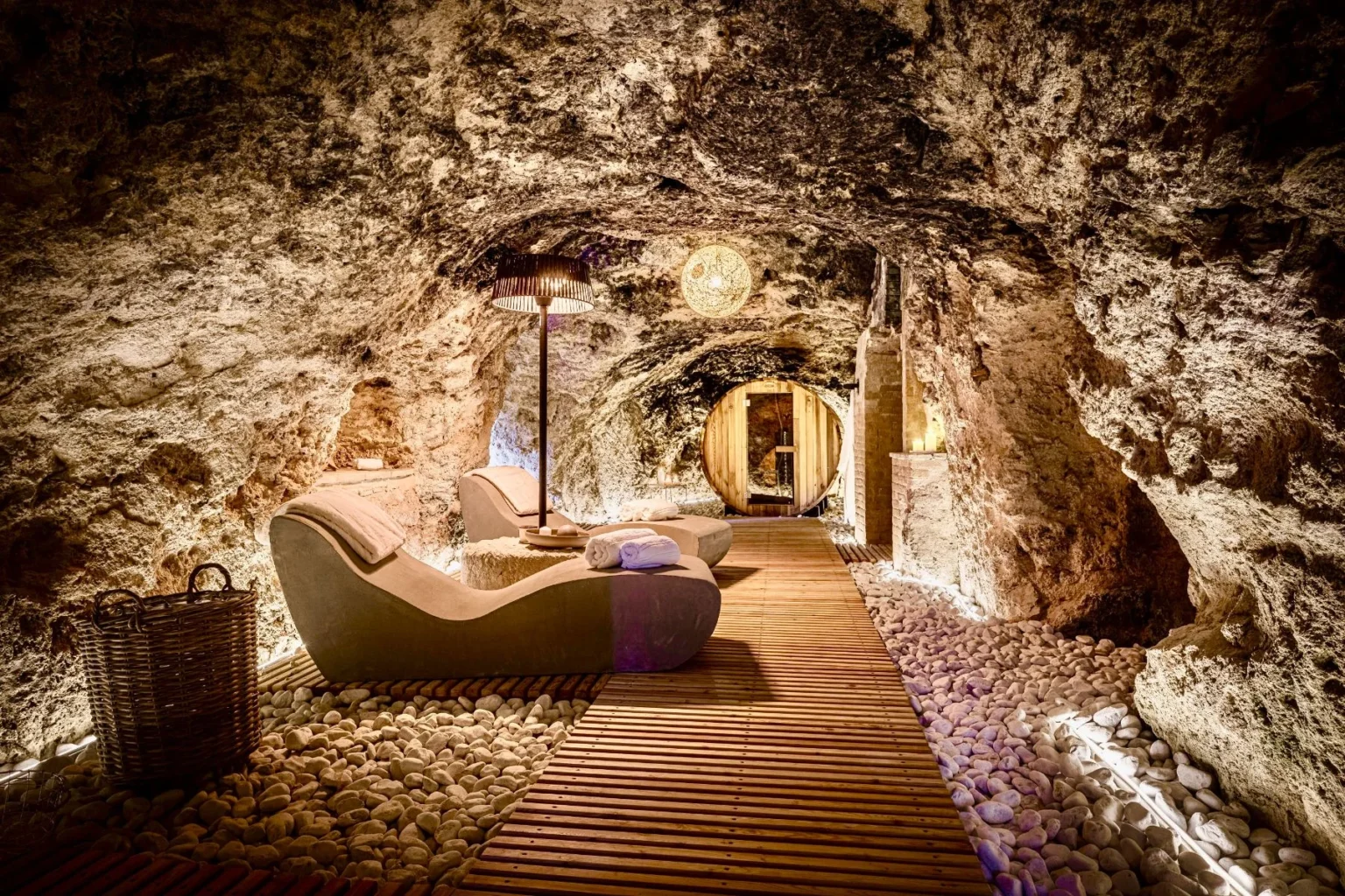 Cozy underground spa featuring smooth stone walls, wooden flooring, and modern lounge chairs, illuminated by soft lighting and a decorative lamp, creating a serene atmosphere for relaxation and wellness. | Only on SopranoVillas