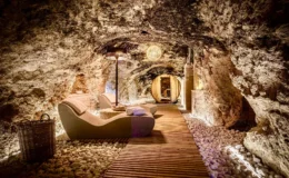Cozy underground spa featuring smooth stone walls, wooden flooring, and modern lounge chairs, illuminated by soft lighting and a decorative lamp, creating a serene atmosphere for relaxation and wellness.