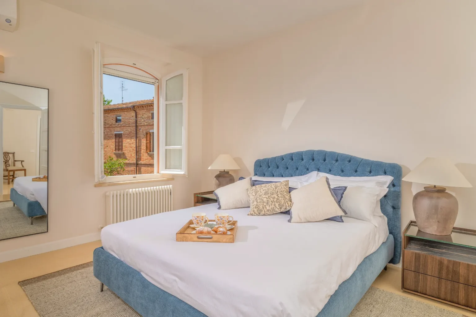 Bright and airy bedroom featuring a cozy blue tufted bed with decorative pillows, a wooden nightstand with a lamp, and a tray of pastries on the bed. A large window offers a view of a brick building outside, enhancing the room's inviting atmosphere. | Only on SopranoVillas