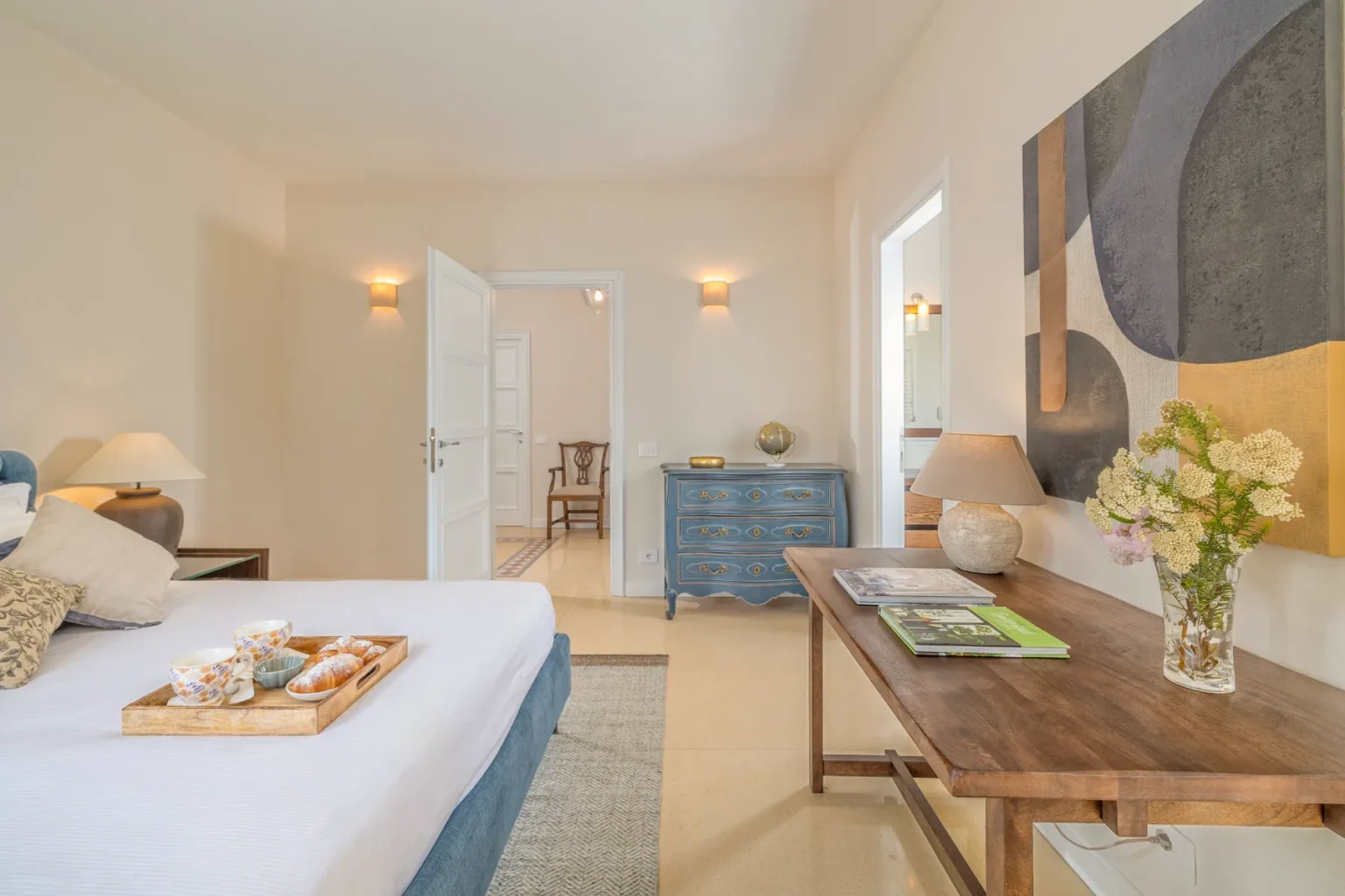 Bright and airy bedroom featuring a comfortable bed with decorative pillows, a wooden tray with breakfast items, and stylish furnishings. The room includes a blue dresser, a wooden side table with a lamp and flowers, and modern artwork on the wall, creating a cozy and inviting atmosphere. | Only on SopranoVillas