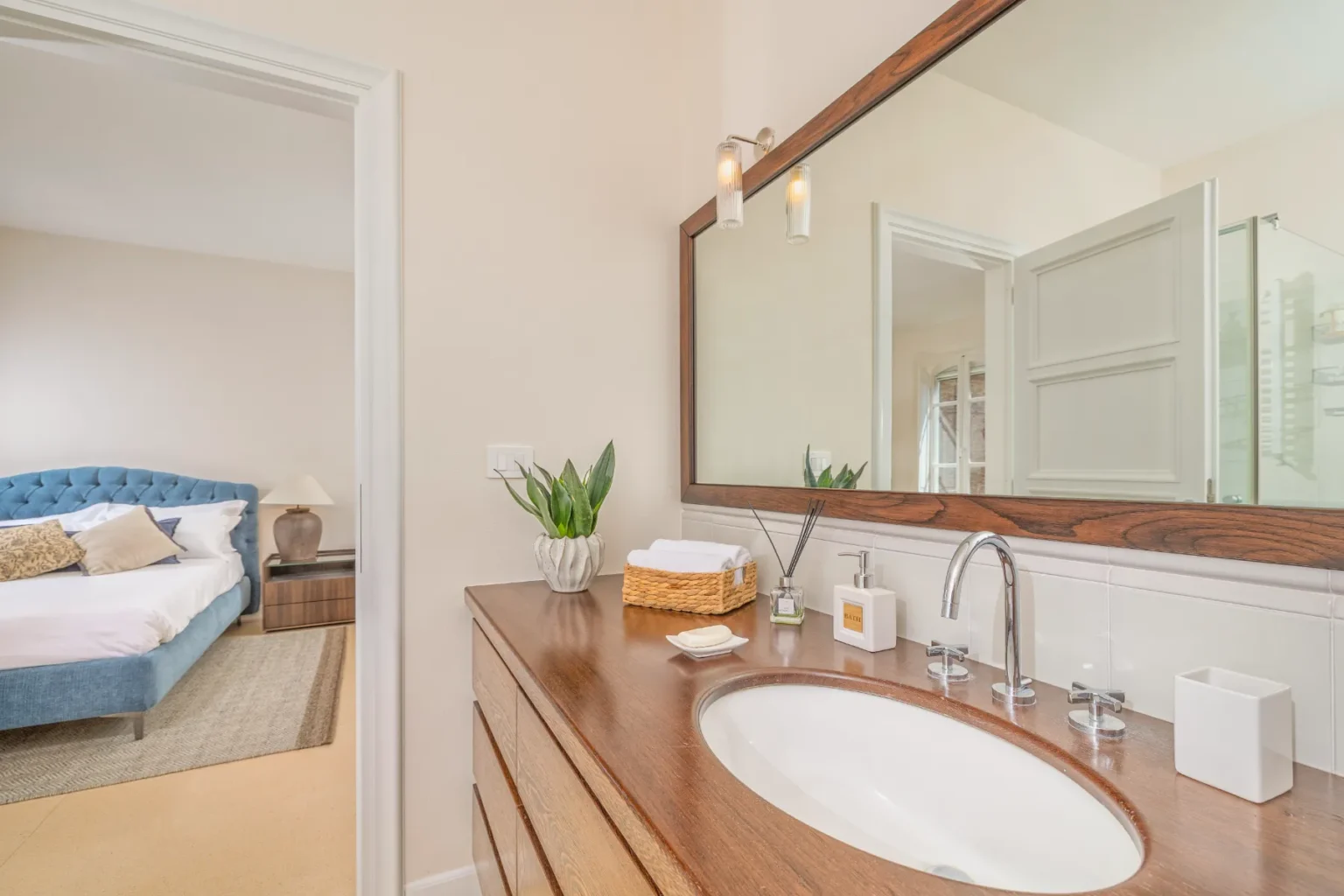 Modern bathroom interior featuring a wooden vanity with a round sink, neatly arranged towels, and decorative plants. A large mirror reflects the light and adds depth to the space, while a cozy bedroom is visible in the background. Ideal for showcasing stylish home decor and interior design elements. | Only on SopranoVillas