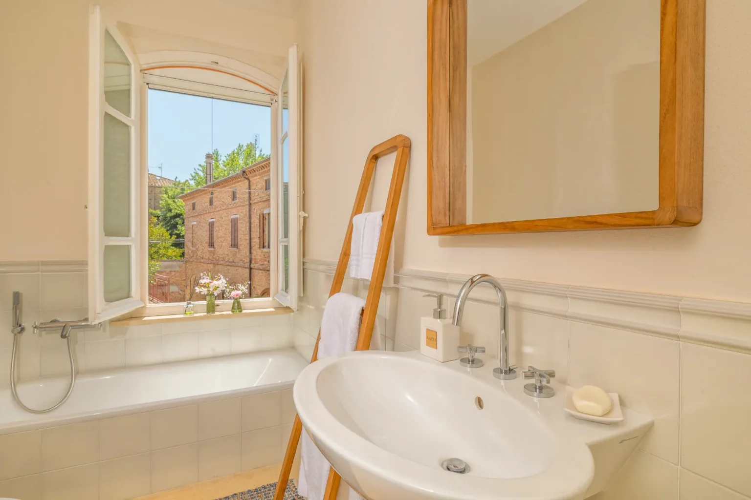 Bright and airy bathroom featuring a large window with a view of a charming exterior, a modern sink with stylish fixtures, a bathtub, and a wooden ladder displaying fresh towels. The neutral color palette and natural light create a serene atmosphere. | Only on SopranoVillas
