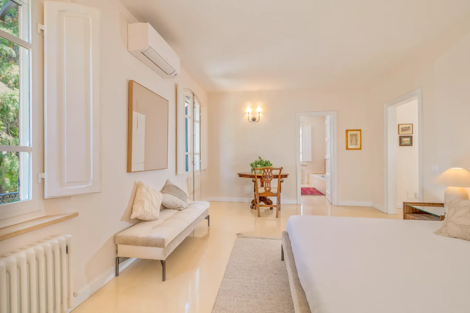 Bright and airy bedroom featuring a comfortable bed, a stylish bench with decorative pillows, and a wooden dining table. Natural light streams in through large windows, complemented by modern air conditioning and elegant wall decor. The room's neutral color palette creates a serene atmosphere, ideal for relaxation. | Only on SopranoVillas