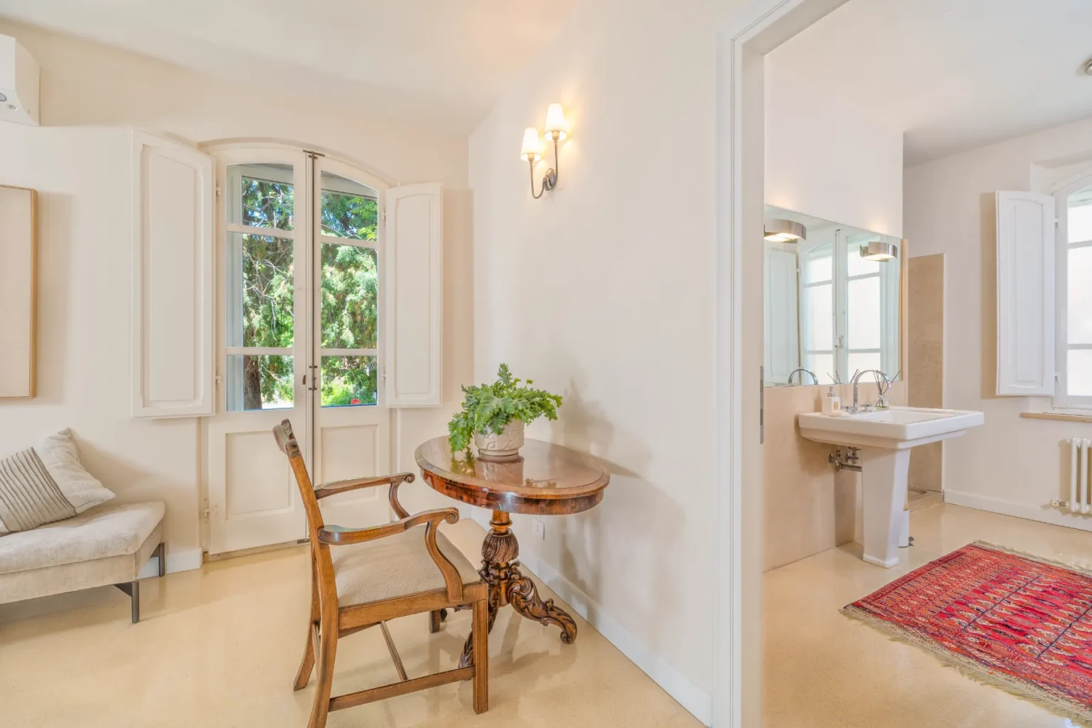 Bright and airy interior featuring a vintage wooden chair and a round table with a potted plant, complemented by large windows that provide natural light and a view of greenery. A modern bathroom is visible in the background, showcasing a stylish sink and elegant fixtures, with a decorative rug on the floor. | Only on SopranoVillas