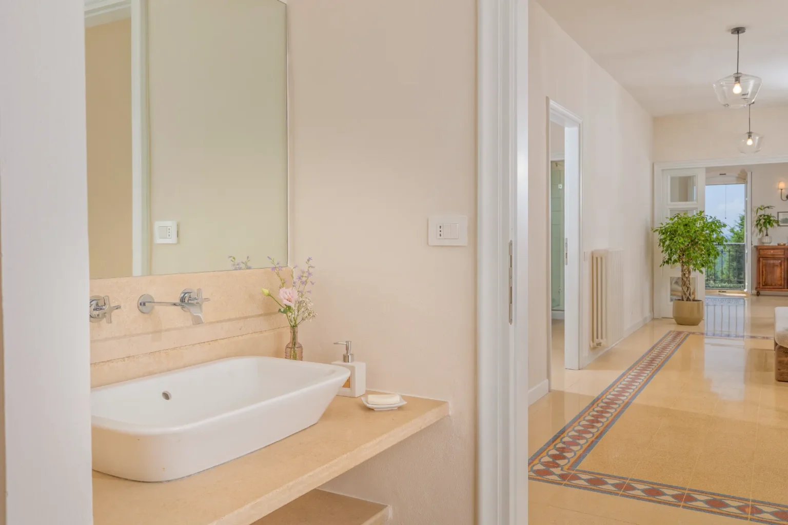 Modern bathroom with a minimalist design featuring a white sink, a large mirror, and a decorative vase with flowers. The neutral color palette and natural light create a serene atmosphere, while the tiled flooring adds a touch of elegance. A glimpse of the adjoining hallway and living area can be seen in the background. | Only on SopranoVillas