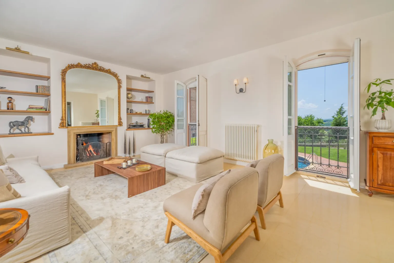 Bright and airy living room featuring a cozy fireplace, elegant mirror, and modern furnishings. The space includes large windows opening to a scenic view, stylish shelving, and a warm color palette, creating an inviting atmosphere for relaxation and socializing. | Only on SopranoVillas