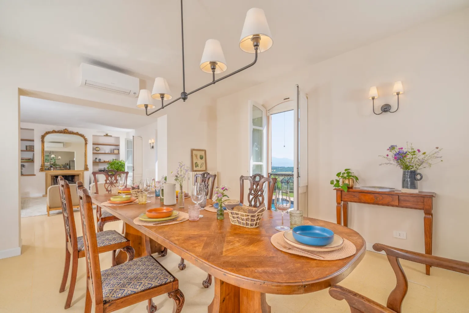 Spacious dining area featuring a wooden table set for a meal, surrounded by elegant chairs and decorative tableware. Natural light floods in through open windows, highlighting the modern light fixtures and tasteful decor, with a view of greenery in the background. | Only on SopranoVillas