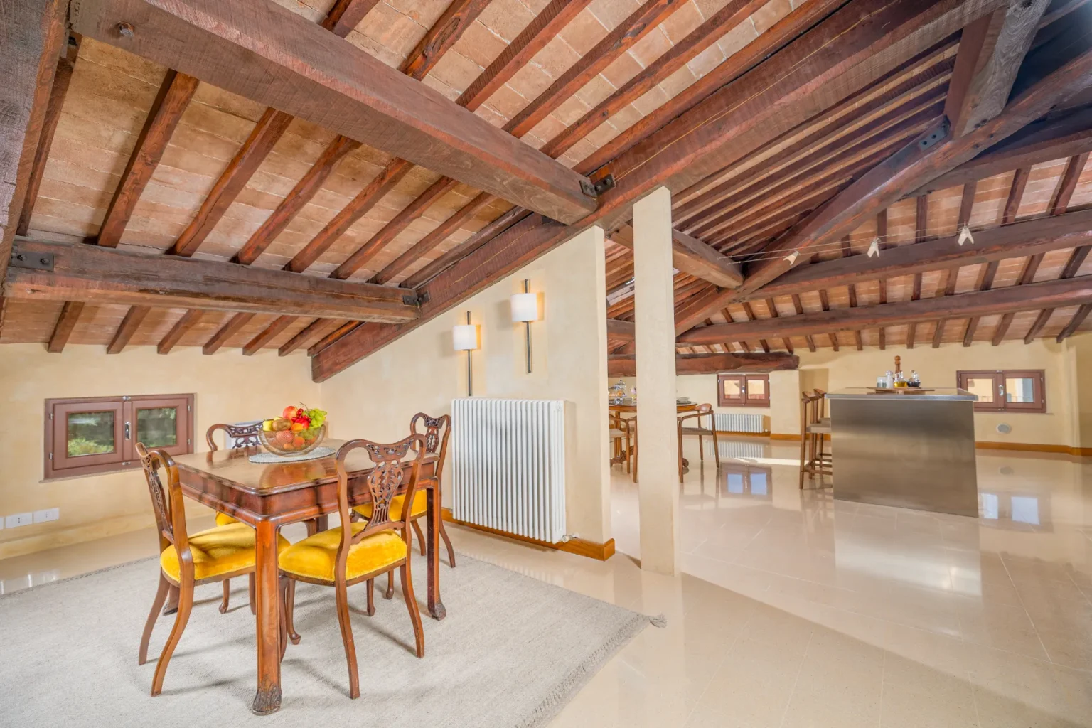 Spacious and stylish attic interior featuring a wooden dining table with yellow upholstered chairs, a fruit centerpiece, and exposed wooden beams. The room includes a modern bar area, elegant lighting, and large windows, creating a warm and inviting atmosphere. | Only on SopranoVillas