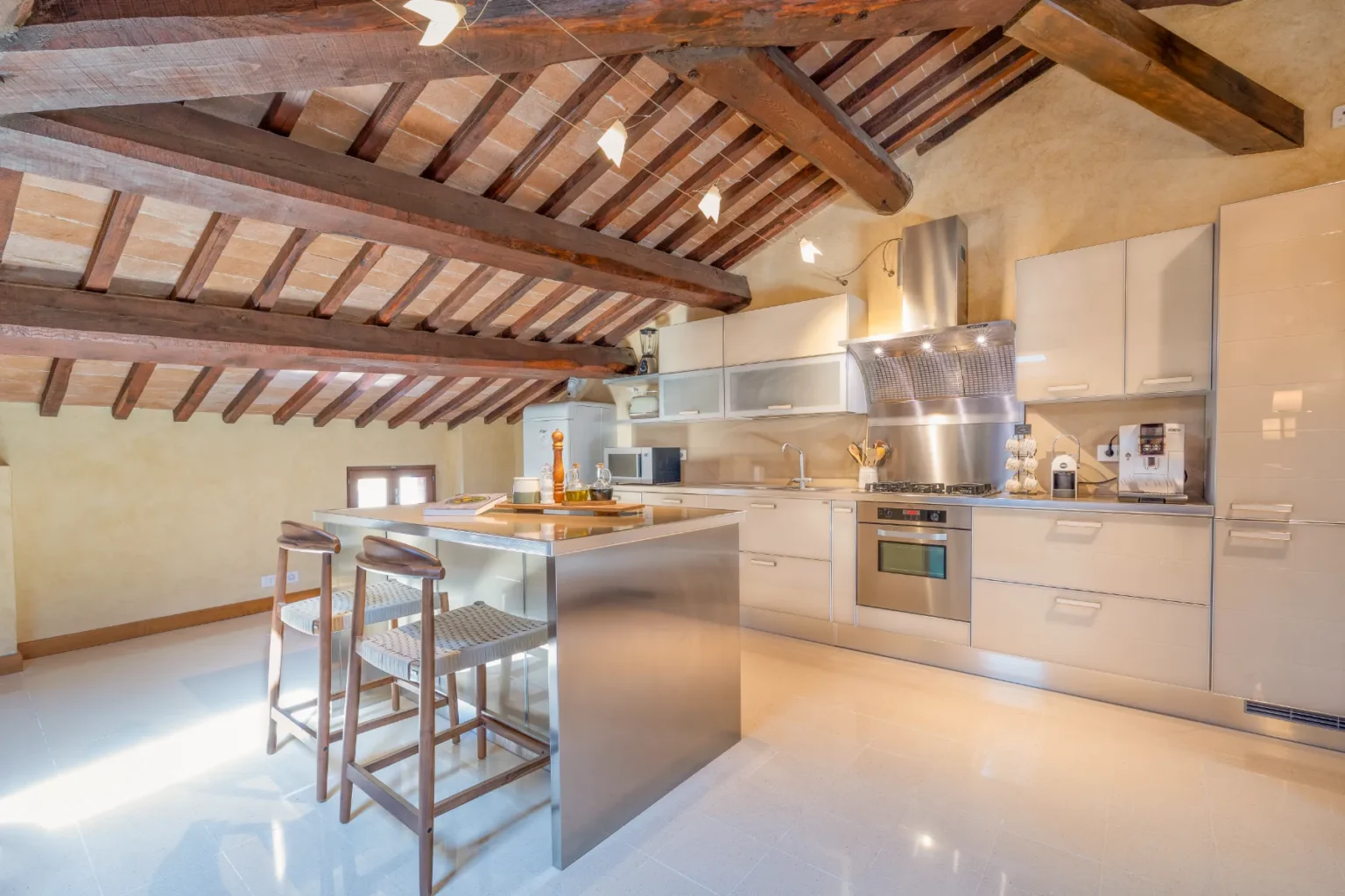 Modern kitchen with exposed wooden beams, sleek cabinetry, and a central island with seating, featuring stainless steel appliances and bright natural light. | Only on SopranoVillas