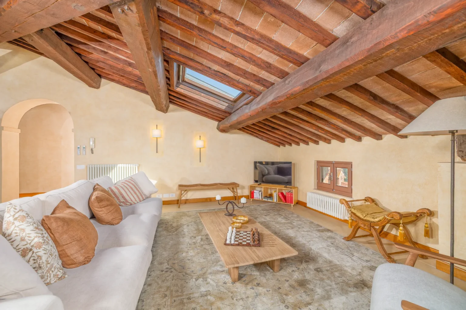 Cozy living room with wooden beams and a skylight, featuring a comfortable white sofa with decorative pillows, a wooden coffee table, a stylish lamp, and a modern TV setup. The warm, neutral tones create an inviting atmosphere, complemented by an elegant area rug. | Only on SopranoVillas