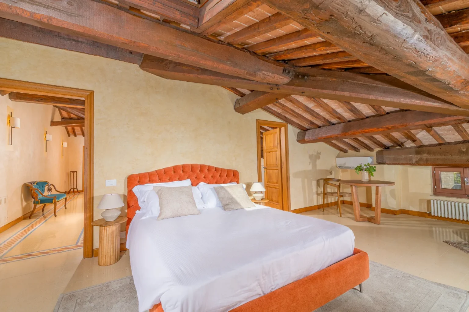 Cozy attic bedroom featuring a plush orange bed with white linens, rustic wooden beams, and a warm color palette. The space includes a small wooden table, decorative pillows, and a stylish armchair in the background, creating an inviting atmosphere. | Only on SopranoVillas