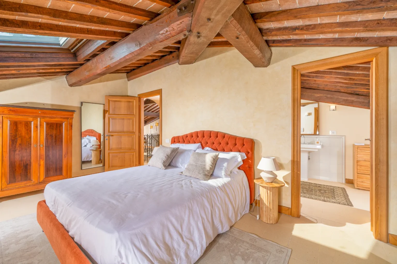 Cozy bedroom featuring a tufted orange bed with decorative pillows, wooden furniture, and exposed beam ceilings. The room includes a large mirror, bedside lamp, and an open doorway leading to a bathroom area, creating a warm and inviting atmosphere. | Only on SopranoVillas