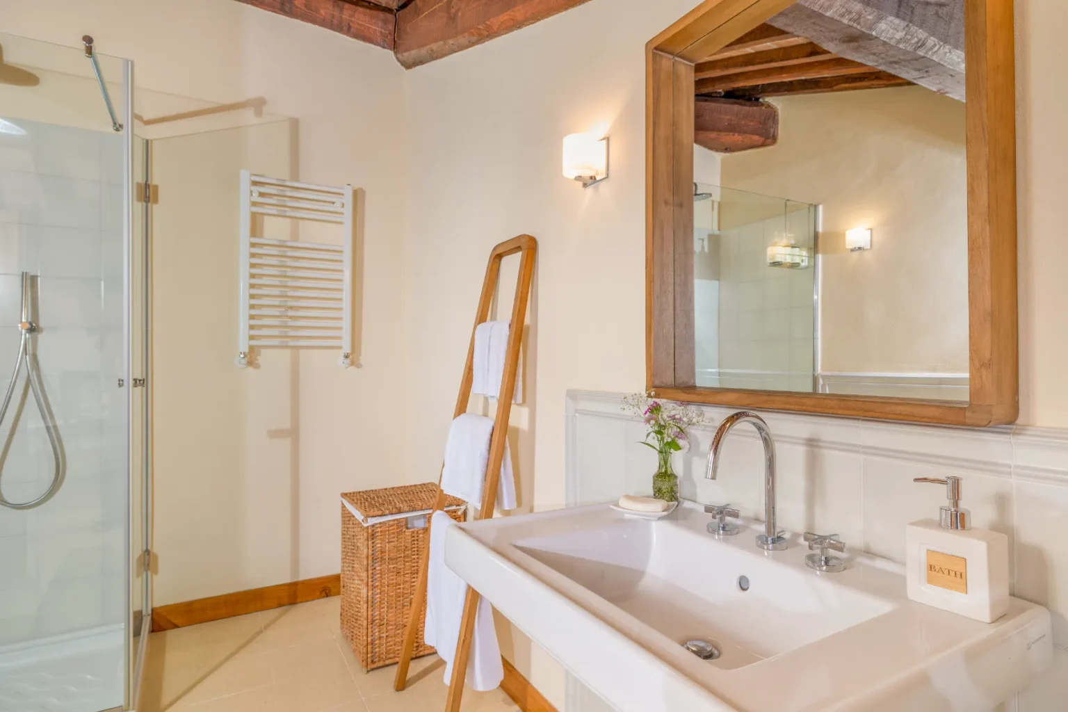 Modern bathroom interior featuring a glass shower, wooden accents, a sleek sink with chrome fixtures, and decorative elements like a flower vase and towel rack. Natural light enhances the warm color palette and inviting atmosphere. | Only on SopranoVillas