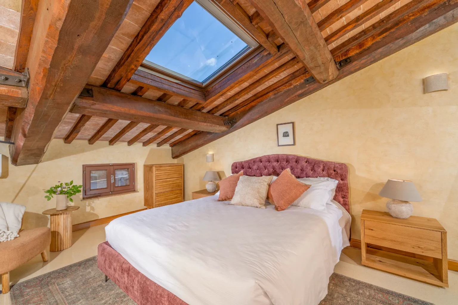 Cozy attic bedroom featuring a plush pink bed with decorative pillows, wooden beams, and a skylight. The room includes a stylish side table, a modern lamp, and a wooden dresser, creating a warm and inviting atmosphere. | Only on SopranoVillas