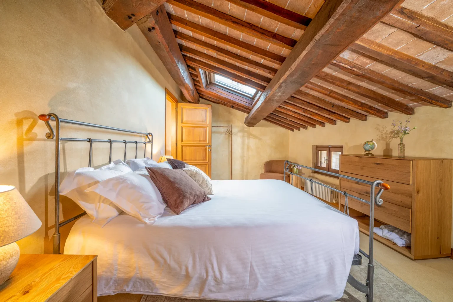 Cozy bedroom with a metal bed frame, plush pillows, and wooden furniture under a rustic slanted ceiling. Features include a skylight, a bedside lamp, and a dresser, creating a warm and inviting atmosphere. Ideal for a comfortable stay. | Only on SopranoVillas