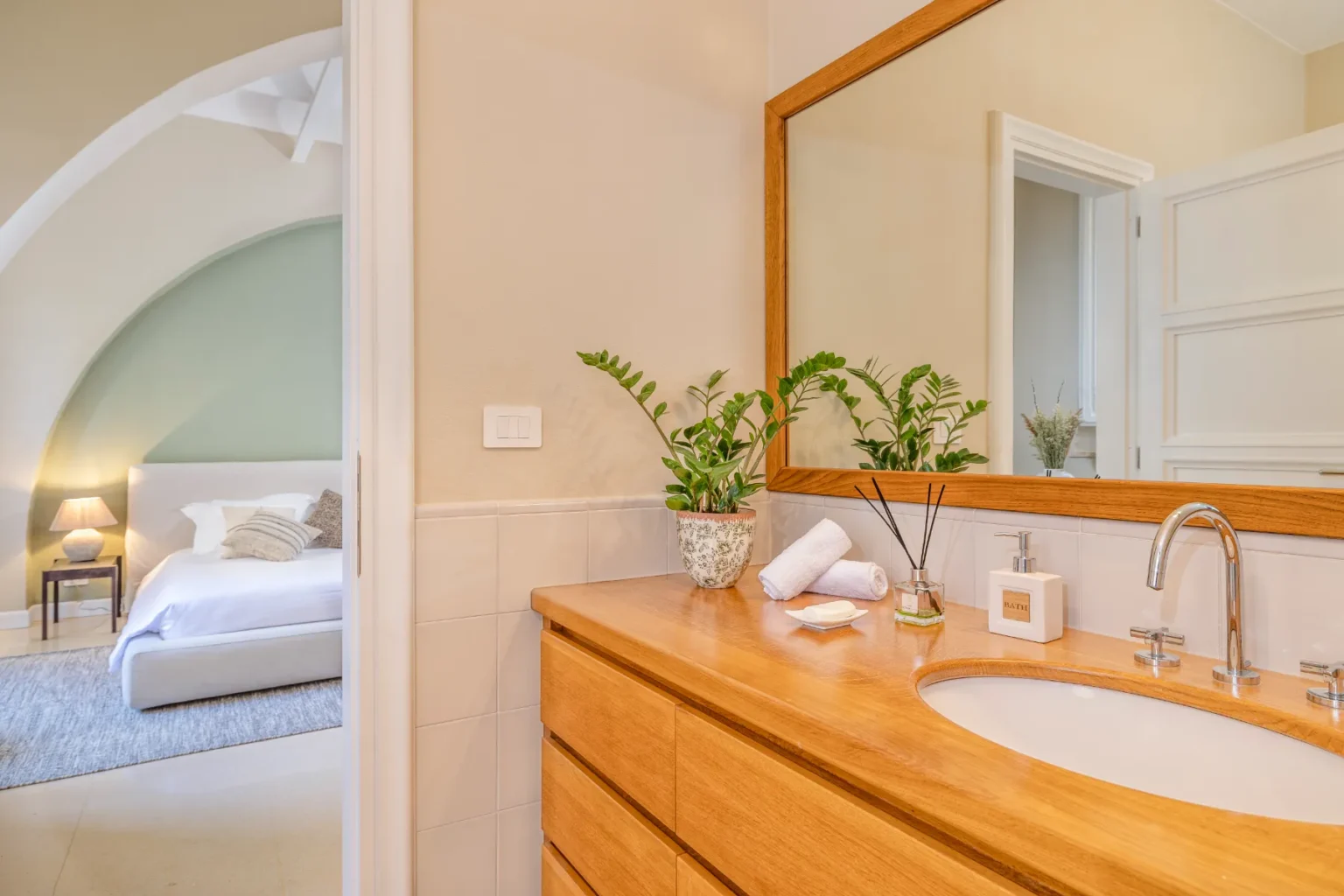 Elegant bathroom interior featuring a wooden vanity with a round sink, decorative plants, and neatly arranged towels. The background reveals a cozy bedroom with soft lighting and a modern bed design, enhancing the serene atmosphere of the space. | Only on SopranoVillas