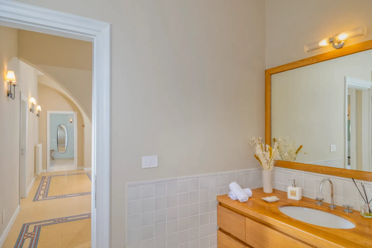 Modern bathroom interior featuring a wooden vanity with a sink, neatly arranged towels, and decorative elements. A large mirror reflects the light from a stylish wall fixture. The view into a hallway showcases elegant lighting and archways, enhancing the spacious feel of the area. | Only on SopranoVillas