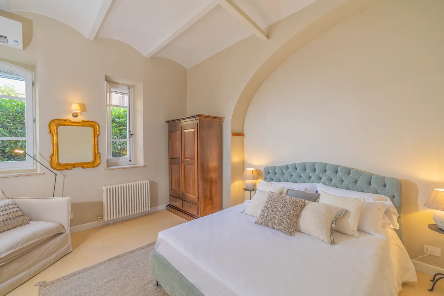 Cozy bedroom featuring a light-colored tufted bed with decorative pillows, a wooden wardrobe, and soft lighting. Large windows provide natural light and views of greenery, while a modern sofa and a stylish gold-framed mirror add elegance to the space. | Only on SopranoVillas