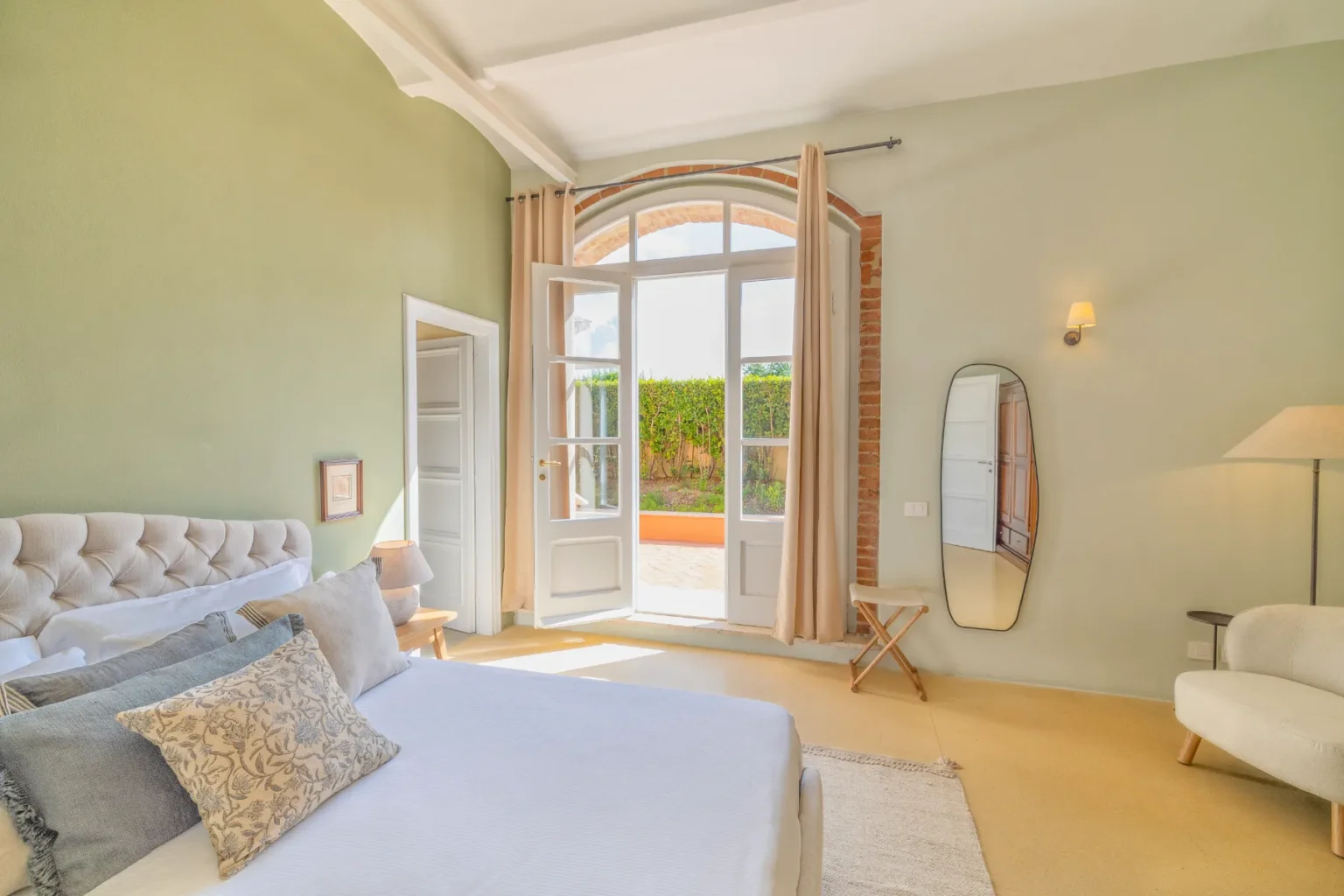 Bright and airy bedroom featuring a tufted headboard, decorative pillows, and large windows with sheer curtains. The room includes a cozy chair, a stylish floor lamp, and a modern mirror, all set against a soothing green wall. Natural light floods the space, highlighting the tranquil atmosphere. | Only on SopranoVillas