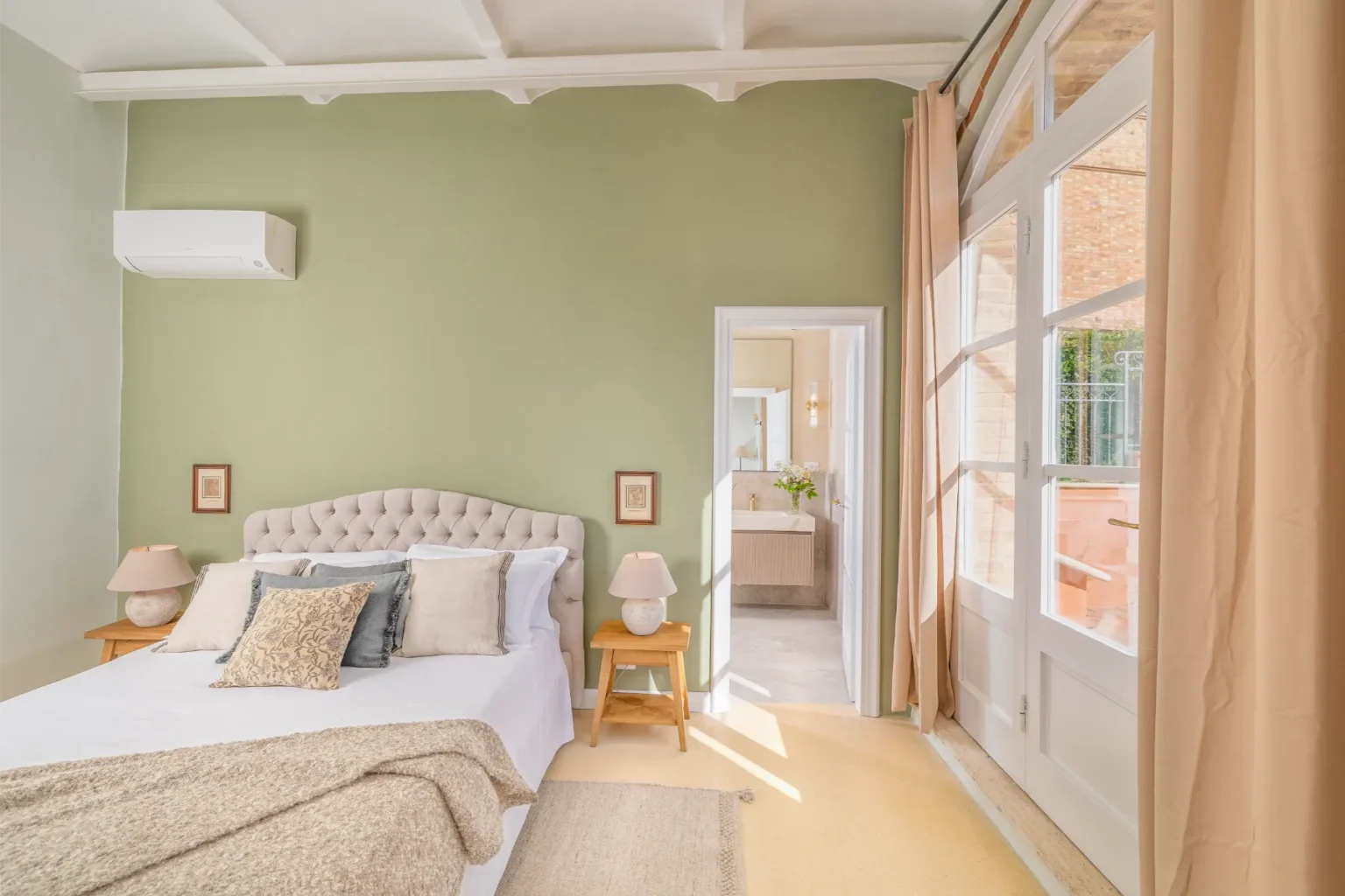 Cozy bedroom with a tufted headboard, decorative pillows, and soft lighting. Features a green accent wall, a bedside lamp, and a doorway leading to a bathroom with natural light and floral decor. Elegant curtains frame large windows, enhancing the room's airy feel. | Only on SopranoVillas