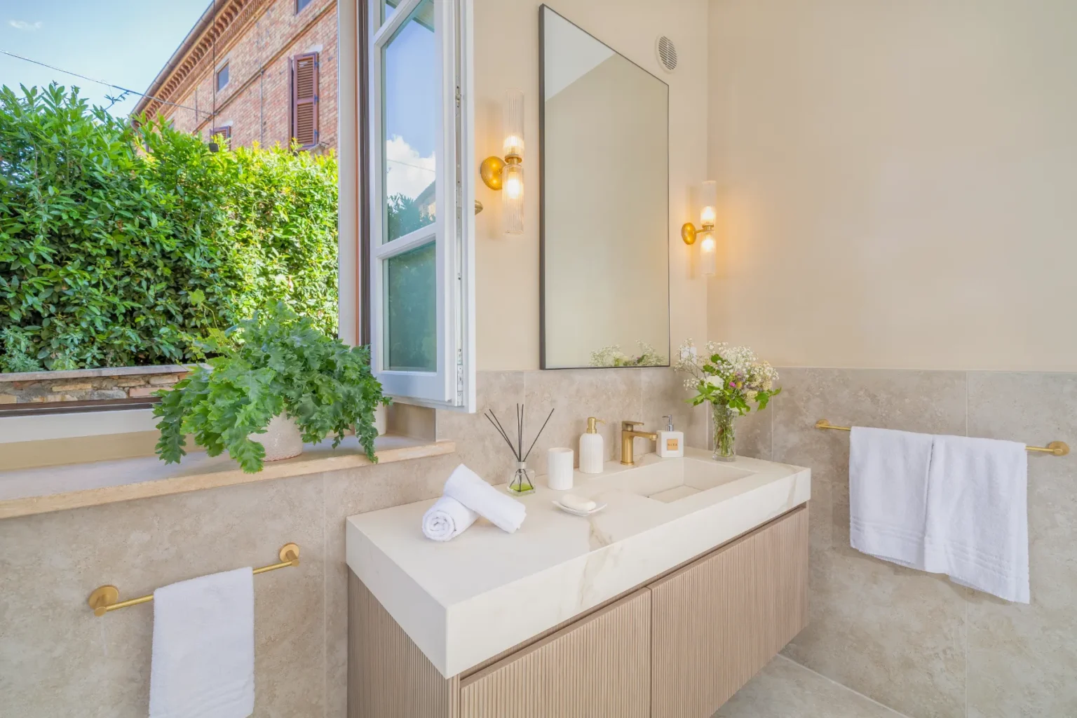 Modern bathroom interior featuring a stylish vanity with a white countertop, wooden cabinetry, and gold fixtures. Natural light streams in through an open window adorned with greenery, enhancing the serene atmosphere. Decorative elements include fresh flowers and neatly arranged towels, contributing to a spa-like ambiance. | Only on SopranoVillas