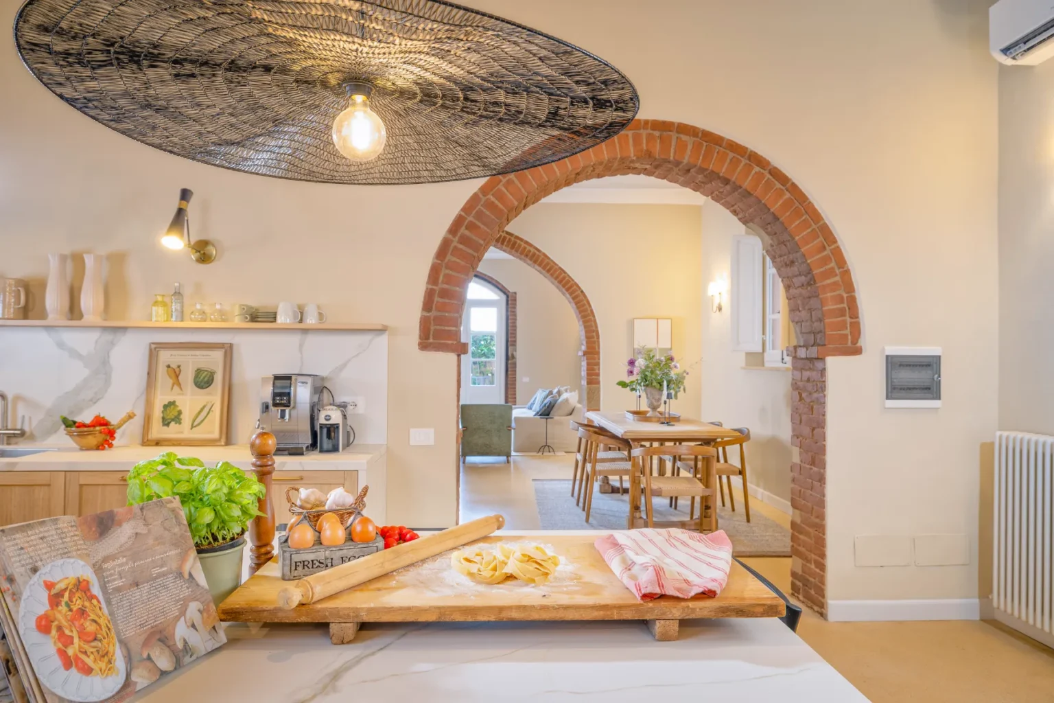Modern kitchen interior featuring a wooden countertop with fresh ingredients, a rolling pin, and a cookbook. The space includes stylish lighting, brick arches, and a dining area visible in the background, creating a warm and inviting atmosphere. | Only on SopranoVillas