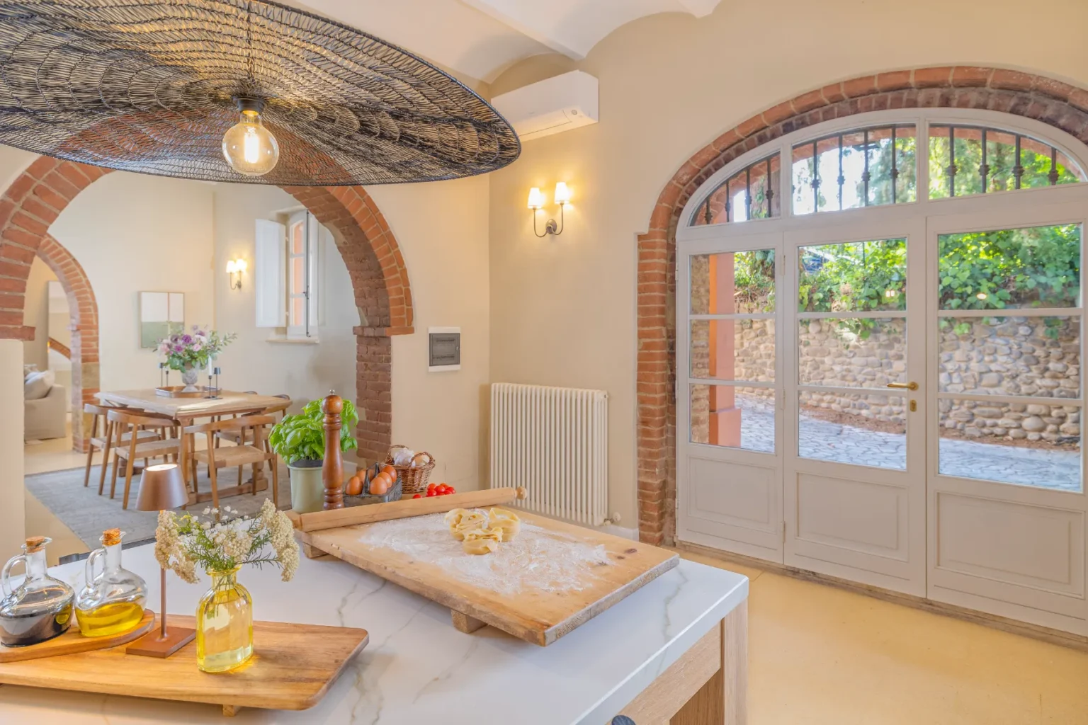 Bright and inviting kitchen interior featuring a marble countertop with fresh ingredients and cooking tools. The space includes a rustic wooden dining table, elegant lighting, and arched brick accents. Large windows open to a scenic outdoor view, enhancing the warm ambiance. | Only on SopranoVillas