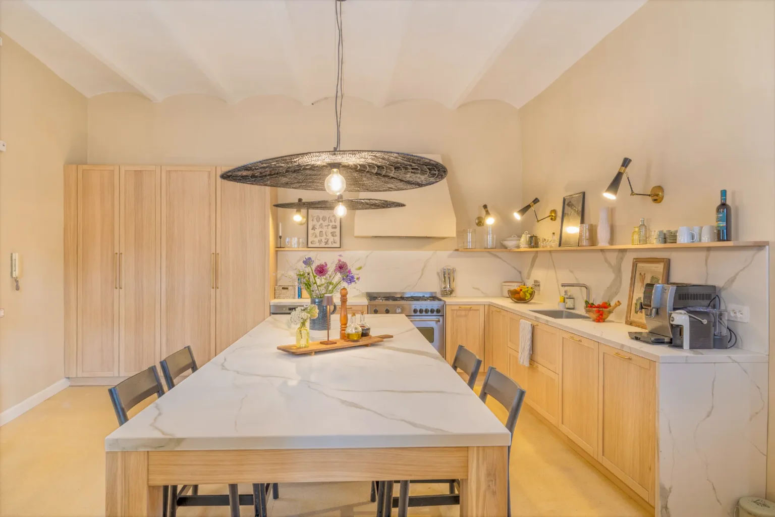 Modern kitchen featuring a large marble island with wooden accents, stylish pendant lighting, and sleek cabinetry. The space includes stainless steel appliances, a coffee machine, and decorative elements like fresh flowers and fruit. | Only on SopranoVillas