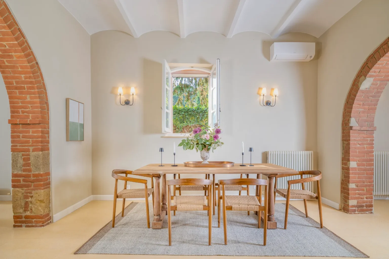 Modern dining room featuring a wooden table surrounded by stylish chairs, illuminated by wall sconces, with a view of greenery through open windows. The space includes decorative elements like a floral centerpiece and a textured rug, complemented by brick archways and neutral wall colors. | Only on SopranoVillas