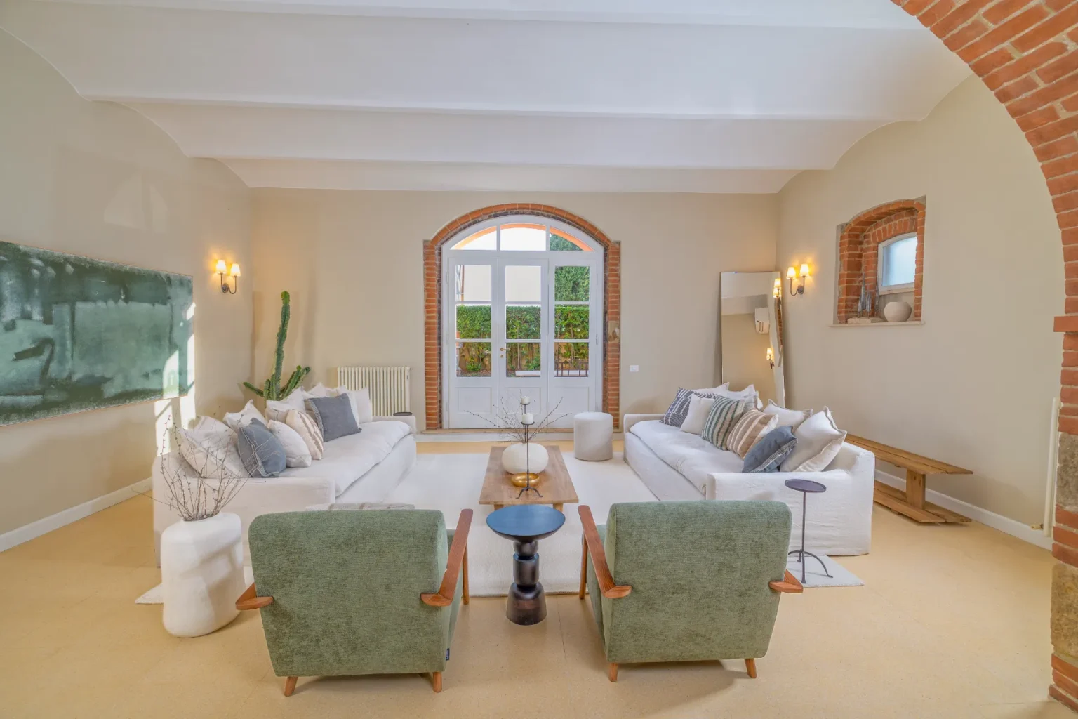 Spacious and modern living room featuring two white sofas adorned with decorative pillows, green armchairs, a wooden coffee table, and a minimalist decor style. Large arched windows allow natural light, complemented by stylish wall sconces and a unique green wall art piece. | Only on SopranoVillas