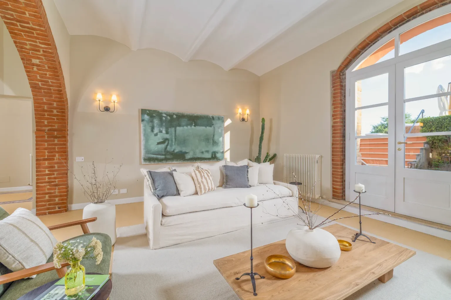 Bright and airy living room featuring a white sofa adorned with decorative pillows, a wooden coffee table with unique home decor, and large windows allowing natural light. The space showcases brick archways, a modern art piece, and green plants, creating a cozy and inviting atmosphere. | Only on SopranoVillas