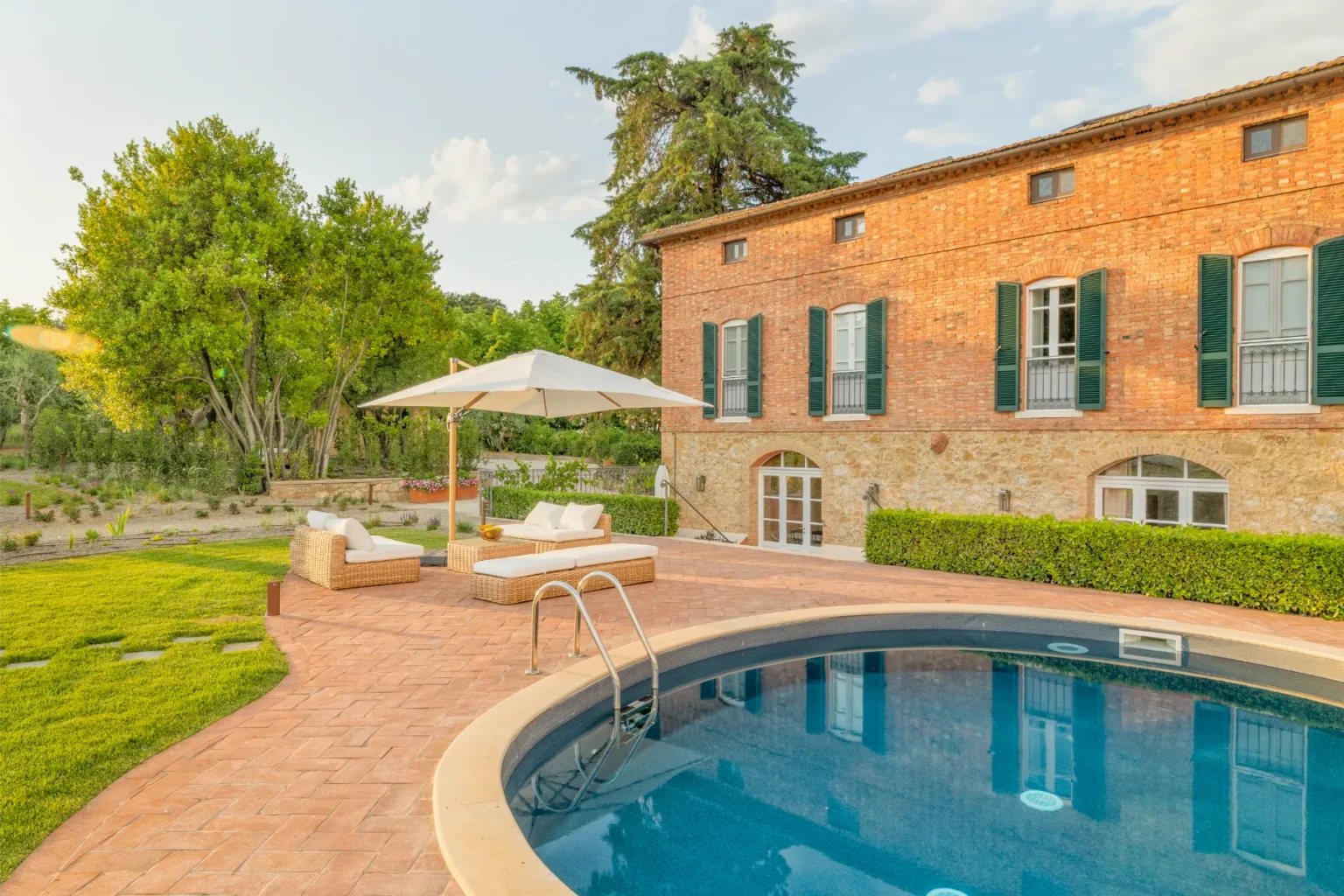 Charming villa with a swimming pool and landscaped garden, featuring a brick facade and green shutters, surrounded by lush trees and outdoor seating areas. Perfect for relaxation and outdoor entertaining. | Only on SopranoVillas