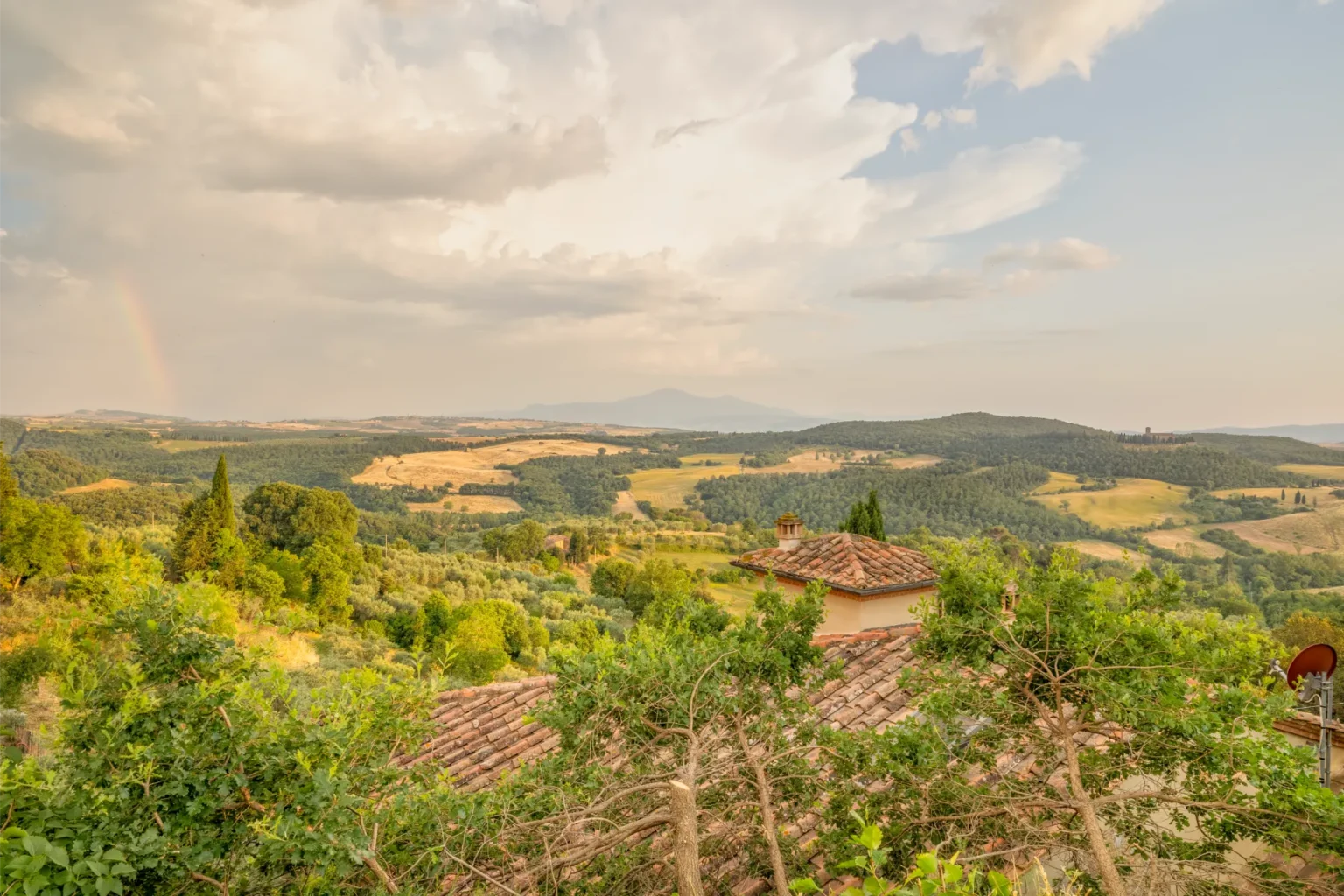 Panoramic view of rolling hills and lush greenery in a Tuscan landscape, featuring a rainbow in the distance and rustic rooftops in the foreground. The scene captures the serene beauty of nature and the characteristic charm of the Italian countryside. | Only on SopranoVillas