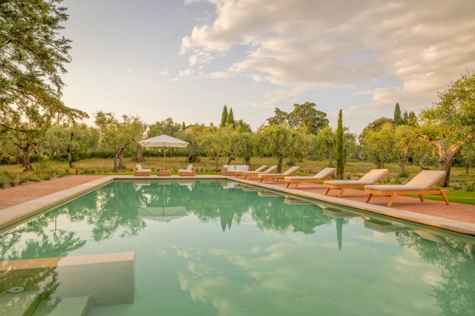 Serene outdoor swimming pool surrounded by olive trees, featuring lounge chairs and a shaded seating area, reflecting a beautiful sky at sunset. Ideal for relaxation and leisure in a tranquil garden setting. | Only on SopranoVillas